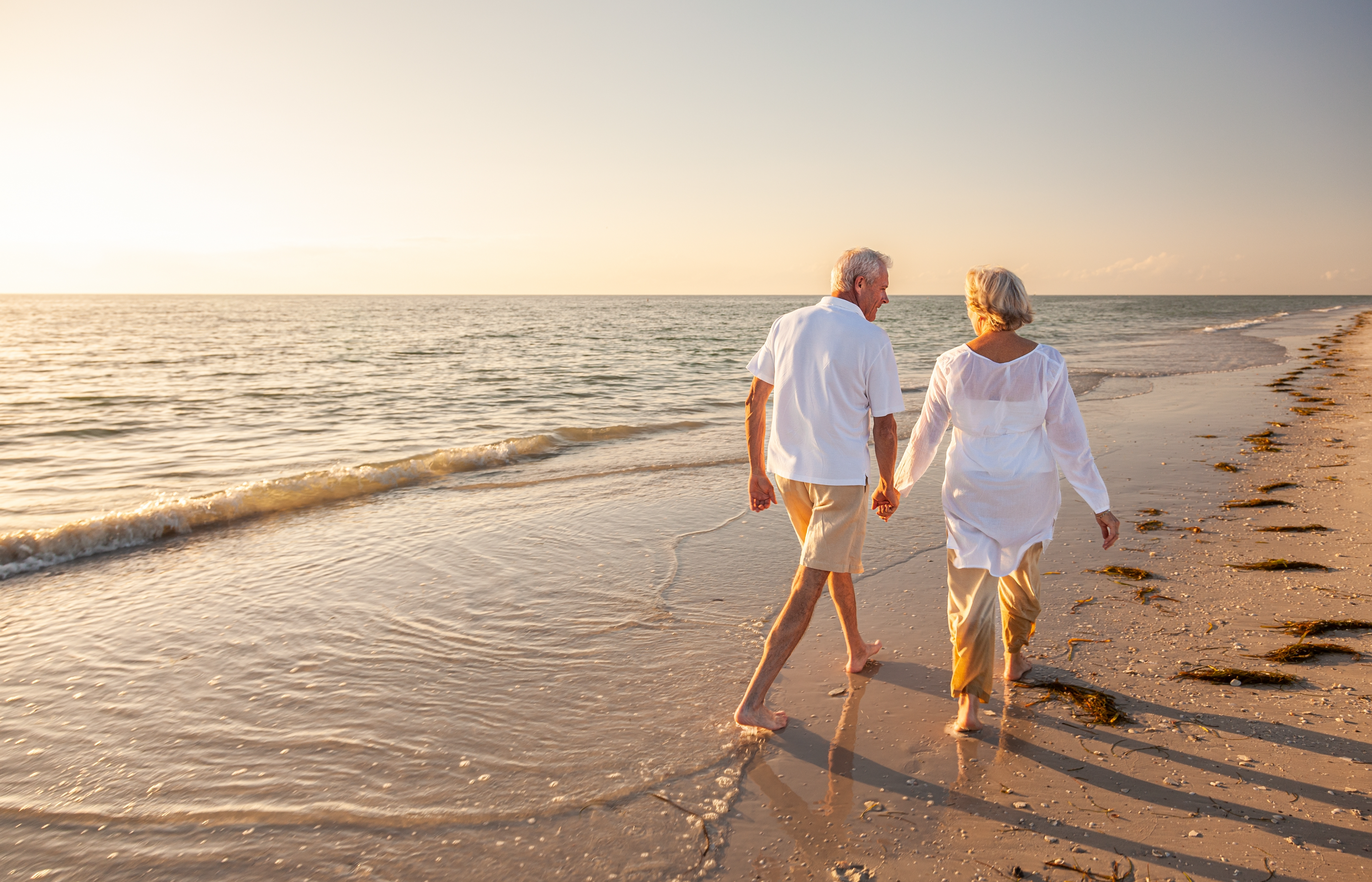 An older couple holding hands walking on the beach during sunset.